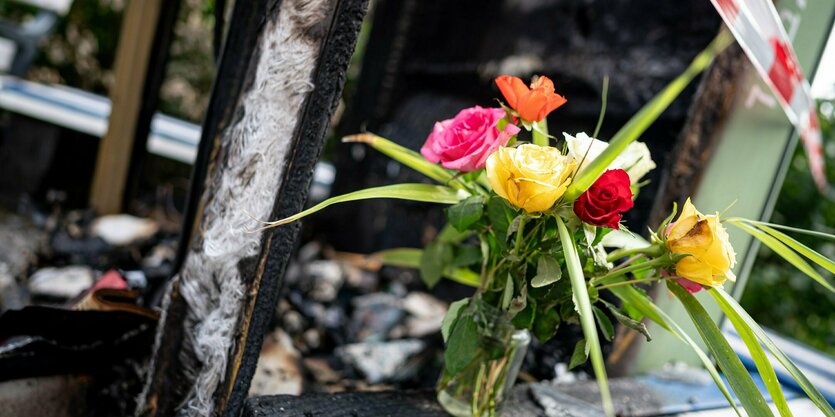 Ein Strauß Rosen steht vor Verkohltem, im Hintergrund ein Absperrband.