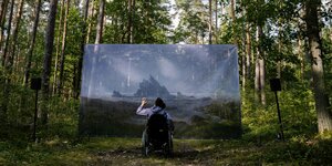 Ein Mann im Rollstuhl schaut auf ein im Wald aufgehängtes Bild