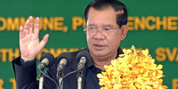 Hun Sen winkt bei seiner letzten offiziellen Rede Anfang August hinter Mikros und Blumen.