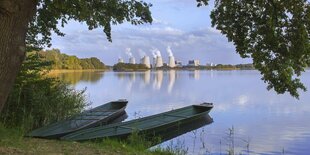 Zwei Boote liegen zwischen Bäumen am Ufer eines Sees, im Hintergrund qualmt das Kohlekraftwerk Jänschwalde.