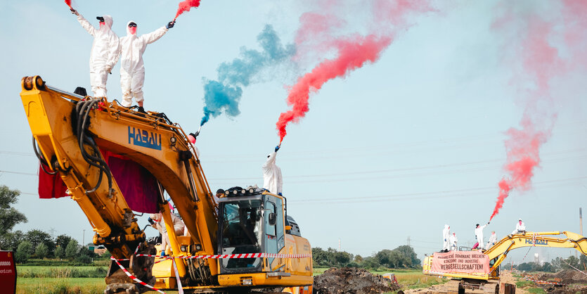 Menschen in weißen Overalls stehen auf Baumaschinen und halten bunte Rauchfackeln in die Luft