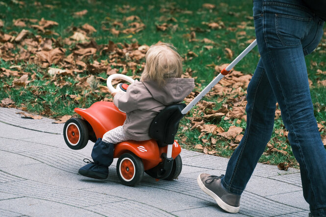 Kind auf einem Bobbycar wird von einer Person geschoben.