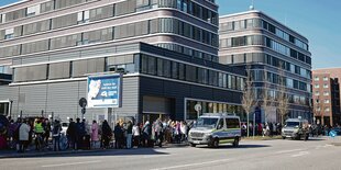 Vor einem grauen Gebäude steht eine lange Schlange wartender Menschen