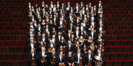 Das Royal Concertgebouworkest Amsterdam steht in einem Publikumssaal inmitten der roten Bestuhlung und schaut nach oben in die Kamera. Die Mitglieder tragen formelle schwarz-weiße bzw. schwarze Kleidung und halten ihre Instrumente in den Händen.