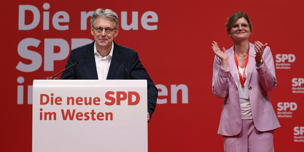 Die neue SPD-NRW-Doppelspitze Achim Post und Sarah Philipp stehen auf einer Bühne am Podium, beide seriös gekleidet