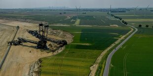 Ein Schaufelbagger gräbt sich in grüne Felder, im Hintergrund Windkraftanlagen