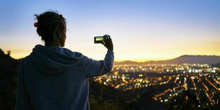 Eine Frau fotografiert mit ihrem Handy eine Stadt von oben in der Dämmerung