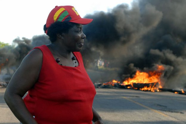 Eine Frau steht auf einer Straße. Im Hintergrund Rauch und Flammen.