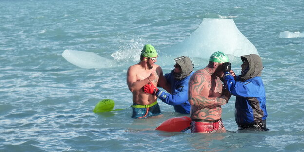 Bibbern im Eissee: Zwei tatowierte Männer stehen mit Badehose im Wasser und werden jeweils von einem Mann in Schutzmontur betreut