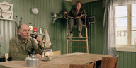 Szene aus dem Film "Kitchen Stories": Links sitzt ein älterer Mann mit Bart an einem Küchentisch. Vor ihm steht ein Wasserkessen und er raucht Pfeife. Im Bildhintergrund sitzt ein Wissenschaftler auf einem Hochhstuhl. Auf seinem Schoß liegt eine Mappe für