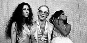 Schwarz-weiss Foto von Cher, Elton John and Diana Ross aus den siebziger Jahren in Glitzeroutfit