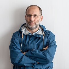 Ira Sachs trägt eine rahmenlose Brille, eine blaue Jacke und hat die Arme verschränkt