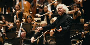 Simon Rattle, ein Mann im schwarzen Anzug und hellen, welligen Haaren, dirigiert das Orchester