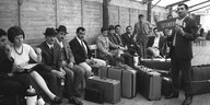 Türkische Gastarbeiter sitzen mit ihren Koffern in einer Wartehalle, vor ihnen steht ein Mann mit einem kleinen Schild: Istanbul