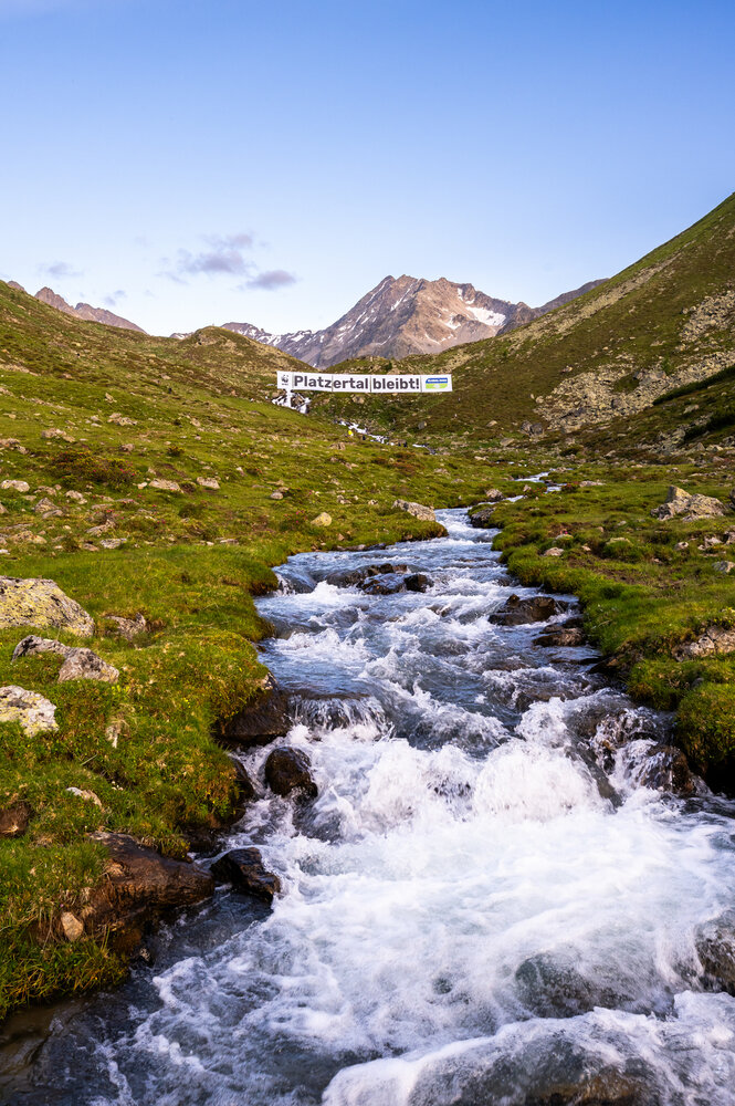 Ein Bach mit kräftigem Wasserlauf, dahinter ein Protestplakat