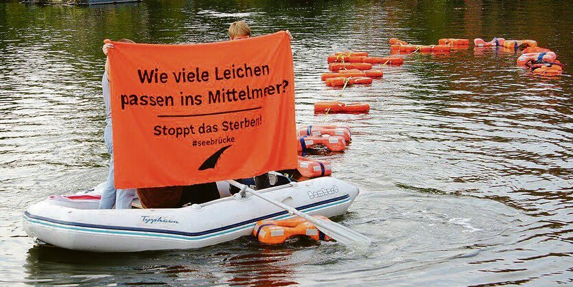 Ein Schlauchboot auf dem Wasser bei einer Protestaktion. Auf einem Transparent steht: "Wie viele Leichen passen ins Mittelmeer? Stoppt das Sterben"