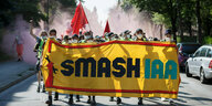 Klimaaktivist:innen laufen in Rauch gehüllt und mit roten Fahnen bestückt eine Straße herunter. Auf dem Fronttransparent steht: "Smash IAA".