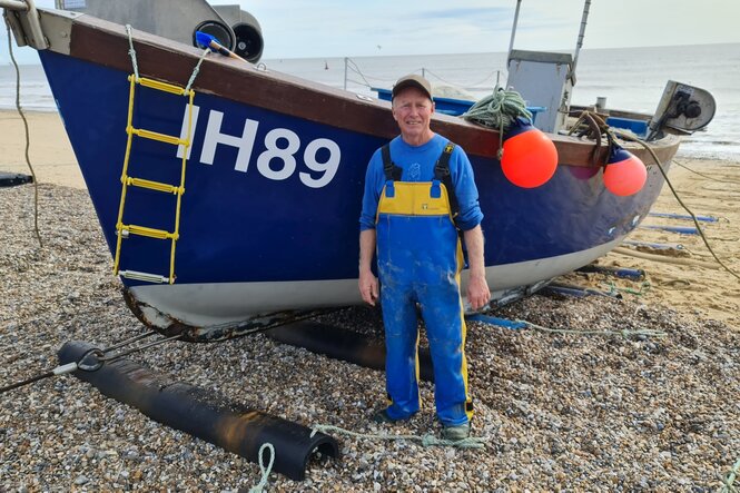 Ein Mann mit blauer Latzträgerhose steht am Strand vpr einem kleinen blauen Boot