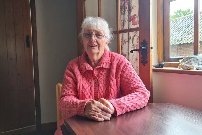 Eine Frau mit grauen Haaren und Brille sitzt an einem Holztisch, auf dem sie die Hände übereinandergefaltet hat