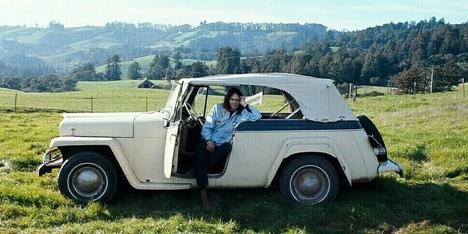 Ein weißer Oldtimer auf einer grünen Wiese, der Himmel ist blau. Auf dem Fahrersitz, die Tür steht offen, sitzt Neil Young