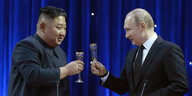 Kim und Putin prosten sich zu vor einem blauen Vorhang mit Sektgläsern