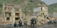 Helfer stehen vor zerstörten Häuser. Hinter den Gebäuden erheben sich kleine Berge