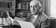 Theodor Adorno sitzt an einem Schreibtisch und hält Notenblätter in den Händen