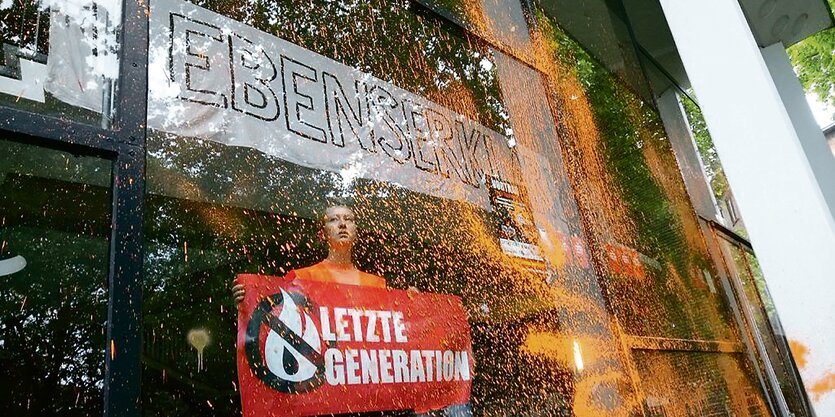 Ein Aktivist mit einem Transparent der "Letzten Generation" steht hinter einer besprühten Fensterscheibe der Uni Hamburg.