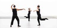 Drei Tänzer:innen in schwarzer Kleidung bewegen sich auf einem weißen Boden. Sie führen eine Hand zum Kopf und halten den anderen Arm horizontal zur Seite gestreckt.