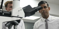Sunak, ein Mann mit dunklen Haar, der ein Hemd und Krawatte trägt, geht zu einem starken Mikroskop