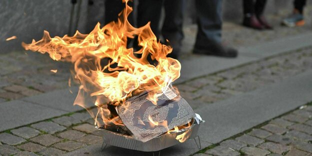Brennender Koran in einer kleinenGrillschale steht aufder Straße, Beine von Männern sind zu sehen