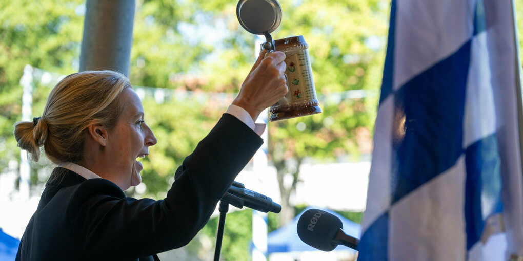 Alice Weidel hält beim Frühschoppen Gillamoos einen Bierkrug hoch, eine bayriscshe Flagge im Vordergrund.