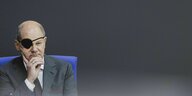 Bundeskanzler Olaf Scholz sitzt auf einem bundestagsblauen Stuhl. Er trägt eine Augenklappe und einen dunklen Anzug. Er hat seine rechte hand ans Kinn gehoben. Der Hintergrund ist dunkelgrau.