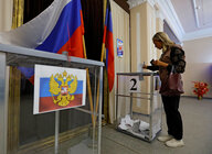 Eine blonde Frau wirft einen Wahlschein in eine Box, im Hintergrund stehen russische Fahnen