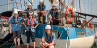 Eine Crew posiert vor einem kleinen hellblau-weißen Segelschiff