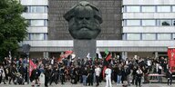 Kundgebung unter einem Karl-Marx-Denkmal. Viele Menschen stehen um das Denkmal mit Flaggen in der Hand.
