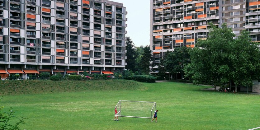 Plattenbauten mit orangfarbenen Sonnensegeln stehen am Rand einer grünen Wiese über die zwei Jungs ein Fussballtor tragen