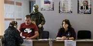 Eine alte Frau mit Kopftuch sitzt an einem Tisch, vor ihr ein Mann, eine Frau, ein vermummter Soldat, ein Plakat mit Putin