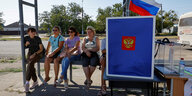 Frauen sitzen auf einer Bank an einer Bushaltestelle, eine russische Wahlurne daneben