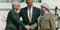 Rabin und Arafat schütteln sich die Hand. Clinton im Anzug steht dahinter und bereitet die Arme aus.