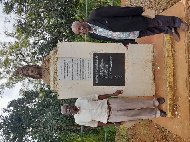 Zablon Kiwelu (links) und Isaria Meli (rechts) stehen am Denkmal für die Hingerichteten in Old Moshi. Das Denkmal ist ein hoher Mamorstein und oben ist ein Kopf.