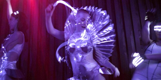 Eine Person in Kostüm tanzt in einem Club. Das Kostüm ist enganliegend und futuristisch. Die Person schwingt eine leuchtende Peitsche. neben der Person rechts und links jeweils ein anderer Mensch.
