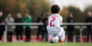 Kleiner Junge von hinten zu sehen, der auf dem Ball sitzt. Im Hintergrund Erwachsene.