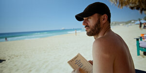 Sebastián Silva sitzt mit Baseballcap und einem Buch von Cioran am Strand.