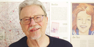 Porträt von Klaus Kordon, Brille kurzer heller Bart, in der Wohnung vor Wand mit Kinderbuchpostern und Stadtplan