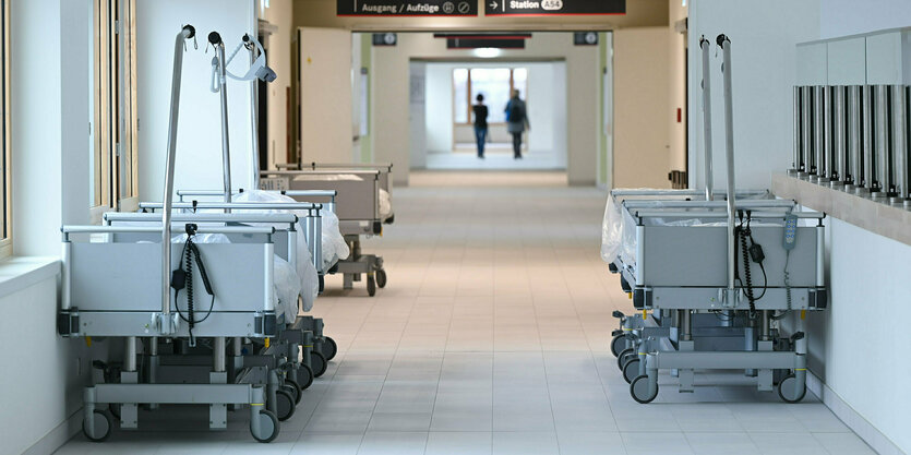 Mehrere Krankenbetten stehen im Flur eines Krankenhauses, am Ende des Flurs sind zwei Personen undeutlich zu erkennen