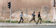 Drei Männer versuchen sich in Sarajevo vor Scharfschützen in Sicherheit zu bringen, hinter ihnen eine Wand mit der Aufschrift: "Welcome to hell"