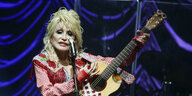 Dolly Parton, eine Frau mit langen blonden Haaren und pinken Fingernägeln, mit Gitarre