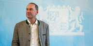 Hubert Aiwanger trägt eine Trachtenjacke, schaut skeptisch zur Seite. Im Hintergrund eine hellblaue Wand mit dem weissen Logo Bayerns