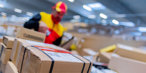 Ein DHL Mitarbeiter steht vor einem Sortierband mit zahlreichen Paketen unterschiedlicher Größe.